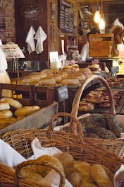 A rustic bakery setting with various types of bread and bagels displayed in wooden crates and wicker baskets. The counter is lined with different baked goods, and the background features a chalkboard menu. Warm lighting adds to the cozy atmosphere, with light bulbs hanging from the ceiling. A person can be seen working behind the counter, surrounded by shelves stocked with books and other items.