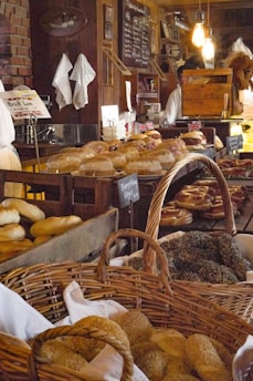 A rustic bakery setting with various types of bread and bagels displayed in wooden crates and wicker baskets. The counter is lined with different baked goods, and the background features a chalkboard menu. Warm lighting adds to the cozy atmosphere, with light bulbs hanging from the ceiling. A person can be seen working behind the counter, surrounded by shelves stocked with books and other items.