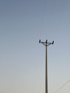 A utility pole stands against a clear sky with several power lines extending horizontally from it. The sky transitions from a light blue at the top to a subtle gradient near the horizon.