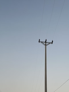 A utility pole stands against a clear sky with several power lines extending horizontally from it. The sky transitions from a light blue at the top to a subtle gradient near the horizon.