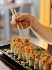 A hand carefully holds chopsticks, picking up a piece of sushi from a rectangular black plate. The sushi is topped with a generous portion of crunchy toppings and garnished with green onions, presenting a colorful and appetizing look.