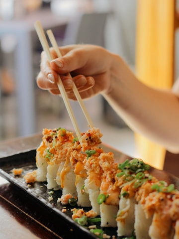 A hand carefully holds chopsticks, picking up a piece of sushi from a rectangular black plate. The sushi is topped with a generous portion of crunchy toppings and garnished with green onions, presenting a colorful and appetizing look.