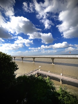 A cloudy sky with patches of cumulus clouds stretches above a wide river. A modern bridge with supporting pillars spans across the river. Lush green trees are visible in the foreground, with a smaller pedestrian bridge crossing a narrower part below.