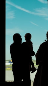 A silhouette of an adult holding a child, standing near a large window at an airport. An airplane is visible on the runway outside against a backdrop of clear blue sky with a few scattered clouds.