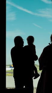 A silhouette of an adult holding a child, standing near a large window at an airport. An airplane is visible on the runway outside against a backdrop of clear blue sky with a few scattered clouds.