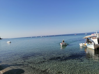 A calm sea with clear blue water stretches into the horizon under a cloudless sky. A few small boats are scattered across the water, with one boat carrying a person paddling closer to the shore. Another larger boat is docked nearby. The scene conveys a peaceful and serene coastal environment.
