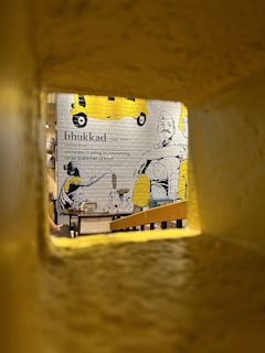 A decorated wall inside a restaurant features a vibrant mural with illustrations of auto rickshaws and a person holding a utensil. The word 'bhukkad' with its definition is prominently displayed. The setting is viewed through a narrow frame, with a warm yellow ambiance.