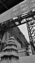 The image depicts a construction site with a large structure covered in scaffolding. In the foreground, there is a decorative pagoda-like structure with intricate designs, set against a backdrop of metal beams and a partially covered building.