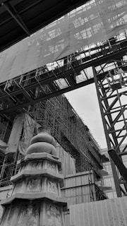 The image depicts a construction site with a large structure covered in scaffolding. In the foreground, there is a decorative pagoda-like structure with intricate designs, set against a backdrop of metal beams and a partially covered building.