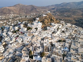 Aerial view of a densely built, traditional Greek town with whitewashed houses clustered around a rocky hill. The town features narrow winding streets, blue-domed roofs, and a backdrop of mountainous terrain.