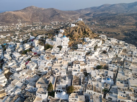 Aerial view of a densely built, traditional Greek town with whitewashed houses clustered around a rocky hill. The town features narrow winding streets, blue-domed roofs, and a backdrop of mountainous terrain.