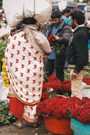 A bustling market scene with a person wearing a floral-patterned garment carrying a large bag on their head. Nearby, vendors are handling and displaying vibrant red roses and other greenery.