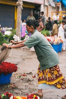 A woman, dressed in a green jacket and a colorful floral skirt, works at an outdoor market. She appears to be sorting or arranging vibrant red roses in a large blue container. The ground is littered with flower petals, and blurred figures of other people and vendors are visible in the background, suggesting a bustling market environment.