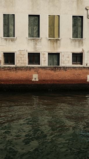 A weathered building facade with closed green and yellow shutters. The lower half features a red brick wall above a calm body of water. The building shows signs of aging with patches of discoloration and some rust on window grilles.