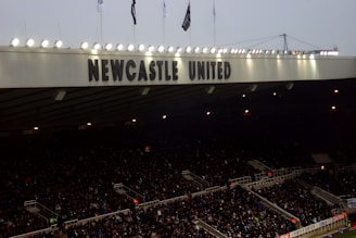 A crowded stadium with many spectators sitting under a large canopy or roof that reads 'NEWCASTLE UNITED' at the top. Bright floodlights illuminate the scene, and banners are visible along the edges. The atmosphere suggests an active event, likely a sports match.