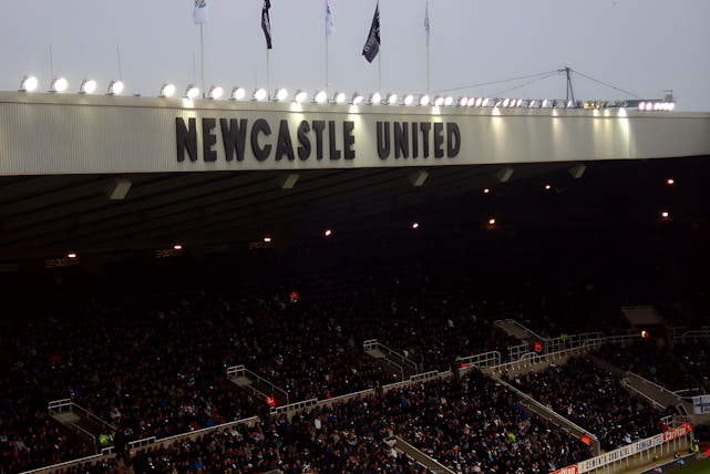 A crowded stadium with many spectators sitting under a large canopy or roof that reads 'NEWCASTLE UNITED' at the top. Bright floodlights illuminate the scene, and banners are visible along the edges. The atmosphere suggests an active event, likely a sports match.
