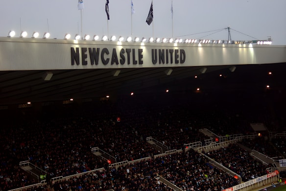 A crowded stadium with many spectators sitting under a large canopy or roof that reads 'NEWCASTLE UNITED' at the top. Bright floodlights illuminate the scene, and banners are visible along the edges. The atmosphere suggests an active event, likely a sports match.