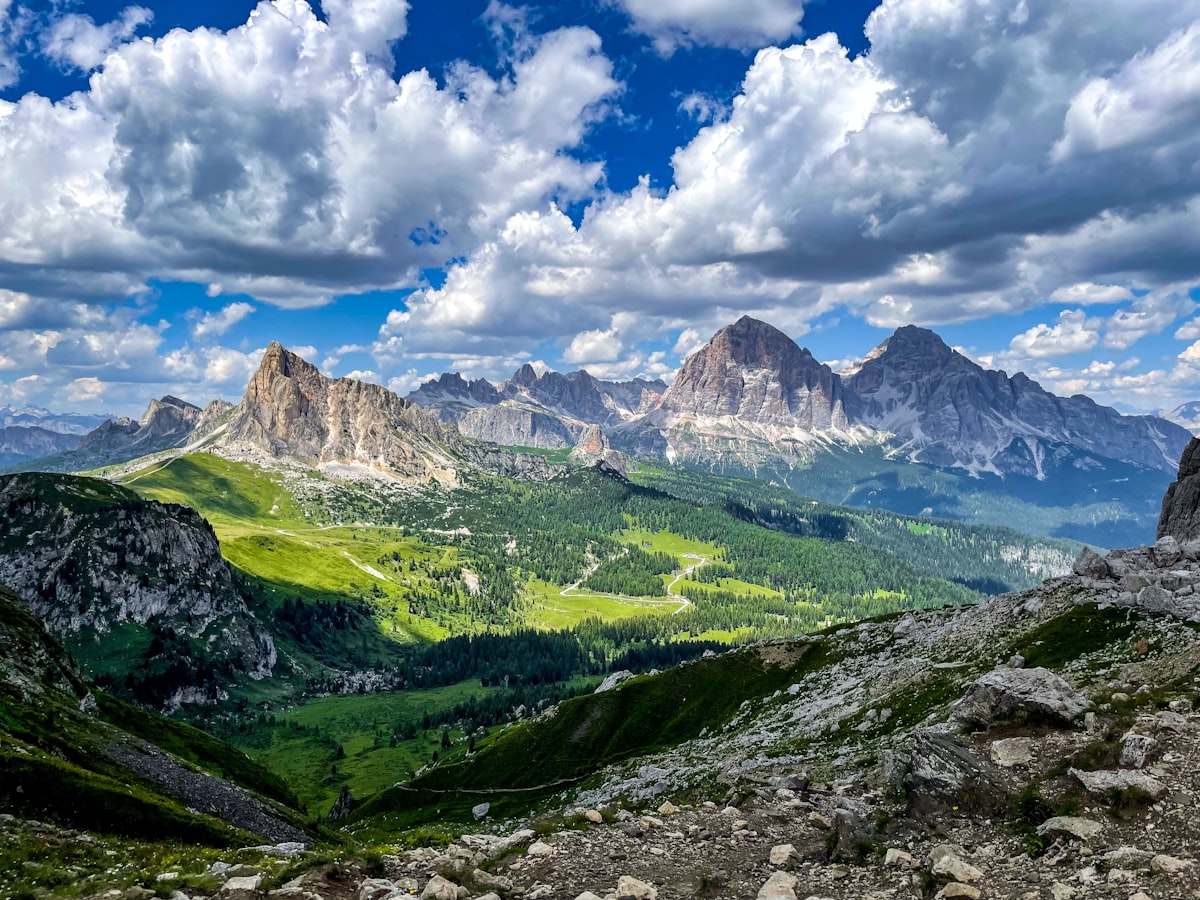 Pale limestone towers rising above green meadows and a winding valley under towering cumulus clouds in the Dolomites