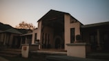A rustic outdoor patio scene featuring a building with a covered entrance and minimalist design. The building has a sloped roof and is surrounded by a peaceful natural setting. There are potted plants on the ledge in the foreground, while outdoor furniture is visible on the patio. The lighting suggests a sunset or sunrise, creating a moody atmosphere.