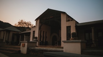 A rustic outdoor patio scene featuring a building with a covered entrance and minimalist design. The building has a sloped roof and is surrounded by a peaceful natural setting. There are potted plants on the ledge in the foreground, while outdoor furniture is visible on the patio. The lighting suggests a sunset or sunrise, creating a moody atmosphere.