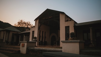 A rustic outdoor patio scene featuring a building with a covered entrance and minimalist design. The building has a sloped roof and is surrounded by a peaceful natural setting. There are potted plants on the ledge in the foreground, while outdoor furniture is visible on the patio. The lighting suggests a sunset or sunrise, creating a moody atmosphere.