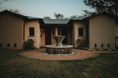 A quaint single-story house with a light-colored exterior and a central circular fountain made of stone in the foreground. The house has a corrugated metal roof and small windows with dark frames. The garden area features well-maintained grass, stone pathways, and a few shrubs and trees surrounding the building.