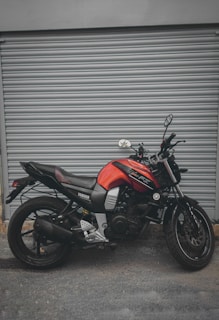 A red and black Yamaha motorcycle is parked in front of a closed gray metal shutter. The bike is positioned side-on, showcasing its sleek design and modern features. The background is plain and industrial, highlighting the motorcycle.