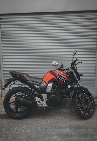 A red and black Yamaha motorcycle is parked in front of a closed gray metal shutter. The bike is positioned side-on, showcasing its sleek design and modern features. The background is plain and industrial, highlighting the motorcycle.