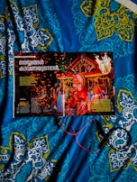 A magazine opened to a page featuring a vibrant traditional celebration with people in colorful attire and a large ornate headdress. The background includes a decorated structure with flames and sparks adding to the festive atmosphere. The image is placed on a blue and patterned quilt, and some red and black wires or cords are seen.