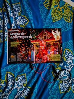 A magazine opened to a page featuring a vibrant traditional celebration with people in colorful attire and a large ornate headdress. The background includes a decorated structure with flames and sparks adding to the festive atmosphere. The image is placed on a blue and patterned quilt, and some red and black wires or cords are seen.