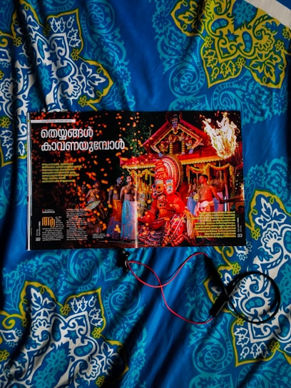 A magazine opened to a page featuring a vibrant traditional celebration with people in colorful attire and a large ornate headdress. The background includes a decorated structure with flames and sparks adding to the festive atmosphere. The image is placed on a blue and patterned quilt, and some red and black wires or cords are seen.