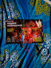 A magazine opened to a page featuring a vibrant traditional celebration with people in colorful attire and a large ornate headdress. The background includes a decorated structure with flames and sparks adding to the festive atmosphere. The image is placed on a blue and patterned quilt, and some red and black wires or cords are seen.