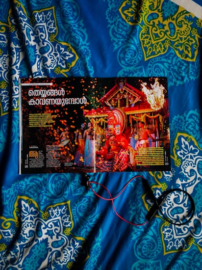 A magazine opened to a page featuring a vibrant traditional celebration with people in colorful attire and a large ornate headdress. The background includes a decorated structure with flames and sparks adding to the festive atmosphere. The image is placed on a blue and patterned quilt, and some red and black wires or cords are seen.