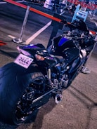 A sleek, black motorcycle is parked in what appears to be an outdoor lot. The bike is angled to showcase its wide rear tire, streamlined body, and a shiny exhaust. A license plate from Qatar is visible. Red and black barriers surround the area, with some chairs and other objects blurred in the background.