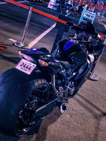 A sleek, black motorcycle is parked in what appears to be an outdoor lot. The bike is angled to showcase its wide rear tire, streamlined body, and a shiny exhaust. A license plate from Qatar is visible. Red and black barriers surround the area, with some chairs and other objects blurred in the background.