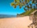 A pristine sandy beach with clear blue skies and tranquil waters. Lush green hills line the background and several small boats are visible near the shore. A few huts made from natural materials are situated on the right, while leafy branches hang above.
