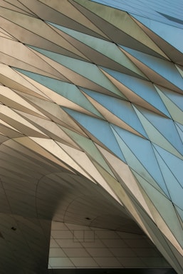 An architectural structure with sharp, angular lines and geometric patterns. The surface consists of sleek, metallic panels that reflect light, creating a textured appearance. The design features overlapping and interlocking triangular shapes.