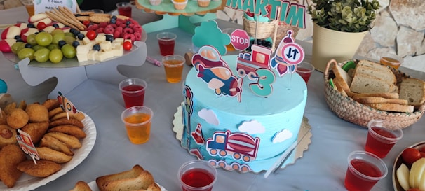 The image features a colorful birthday party setup with a focus on a train-themed birthday cake. The cake is light blue with decorative train cutouts and a '3' indicating the child's age. Surrounding the cake are various treats, including platters of assorted fruits, pastries, and bread. Small plastic cups filled with different colored beverages are scattered around the table. A potted plant adds a touch of greenery to the setting.
