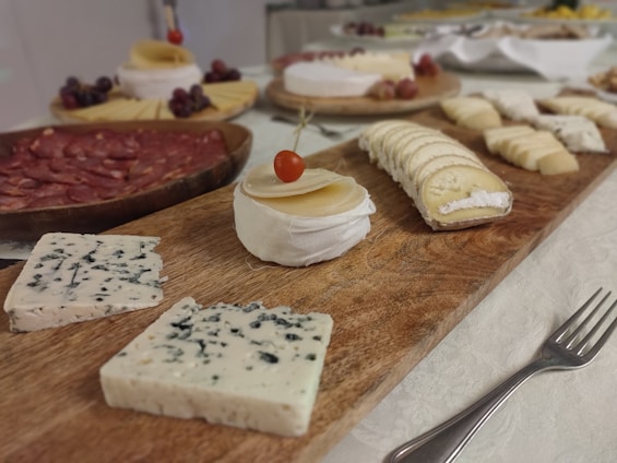 A wooden platter is filled with an assortment of cheeses and cured meats. Various types of cheese are present, including blue cheese, a creamy Brie adorned with a small red fruit on top, and several sliced cheeses. Grapes and salami are also part of the spread, set on separate round wooden boards. The background features additional plates of food, and a metal fork rests on a white tablecloth alongside the main platter.