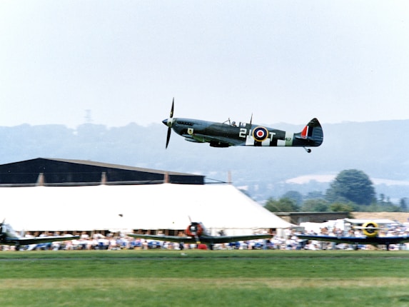 A vintage military aircraft flies low over a grassy field with a background of buildings and spectators. The plane is a Spitfire, identifiable by its distinctive roundel markings and design. The atmosphere suggests an airshow or historical reenactment event.