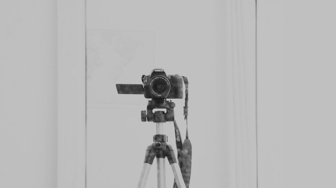 A DSLR camera is mounted on a tripod, positioned indoors in front of a reflective surface. The setup is centrally aligned, with the camera lens facing forward and the camera strap hanging down. The background and surrounding area are minimalistic, emphasizing the photographic equipment.