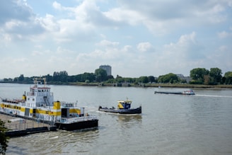 A river scene featuring a large yellow and white ferry docked at the riverside, with trees and buildings visible on the opposite bank. A smaller blue and yellow tugboat is nearby, and a long cargo barge is sailing on the river. The sky is partly cloudy, enhancing the serene atmosphere.