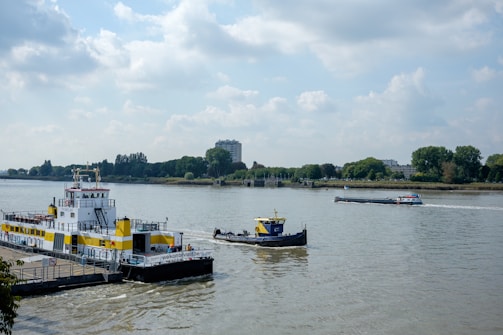 A river scene featuring a large yellow and white ferry docked at the riverside, with trees and buildings visible on the opposite bank. A smaller blue and yellow tugboat is nearby, and a long cargo barge is sailing on the river. The sky is partly cloudy, enhancing the serene atmosphere.