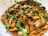 A dish consisting of cooked eggplant pieces mixed with a spicy, crumbly texture, garnished with fresh green cilantro leaves. The mélange is placed in a white bowl, with pieces of brown roasted spices scattered throughout.