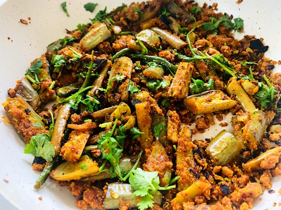 A dish consisting of cooked eggplant pieces mixed with a spicy, crumbly texture, garnished with fresh green cilantro leaves. The m&eacute;lange is placed in a white bowl, with pieces of brown roasted spices scattered throughout.
