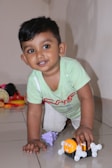 A young child with short dark hair is smiling while kneeling on a shiny tiled floor. The child is wearing a light green shirt with red and white designs and beige checkered pants. They are playing with a small orange toy with white wheels, and there are other colorful toys scattered in the background.