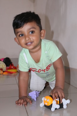 A young child with short dark hair is smiling while kneeling on a shiny tiled floor. The child is wearing a light green shirt with red and white designs and beige checkered pants. They are playing with a small orange toy with white wheels, and there are other colorful toys scattered in the background.