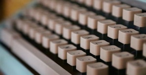 A close-up view of the keys on a vintage typewriter, showcasing rows of cream-colored, square keys with black lettering. The focus is shallow, with the foreground keys clearly visible while the background keys blur gradually.