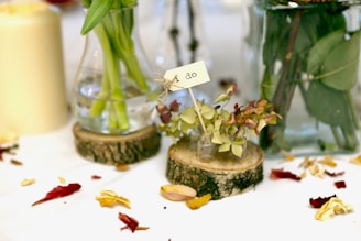 Several glass containers and vases are arranged on a table, each holding flowers and green stems. On the right, a small vial with various leaves and buds is placed on a circular wooden slice, holding a small tag with the text 'I do'. The surface is sprinkled with various colored petals, adding a vibrant touch.