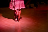 Two individuals are dancing closely on a polished wooden floor, illuminated by soft red lighting. The focus is on their feet; one wearing formal shoes and the other in high heels, suggesting a dance like the tango or waltz.